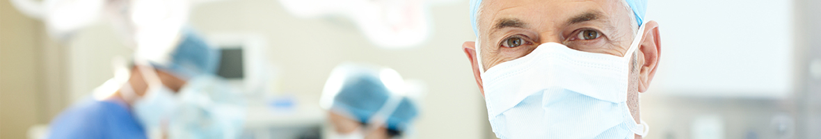 Een zorgverlener van een UMC met een mondkapje kijkt in de camera. Op de achtergrond is een operatiekamer te zien met nog twee UMC-zorgverleners.
